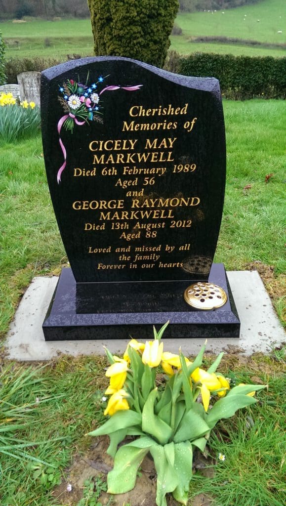 A black granite tombstone with gold inscriptions commemorates Cicely May Markwell and George Raymond Markwell, surrounded by grass. Fresh yellow tulips lie at its base, and the background features a grassy landscape with trees and daffodils.