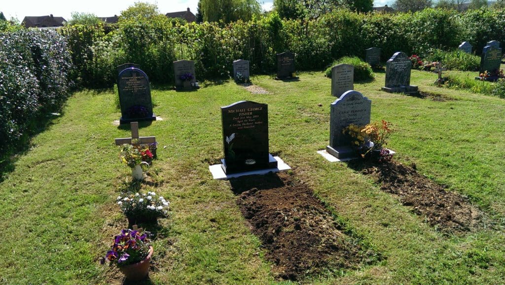 A sunny cemetery scene showing rows of gravestones. Freshly dug earth sits in front of some graves, with flowers and small plants placed around. Lush green grass and dense hedges surround the area.