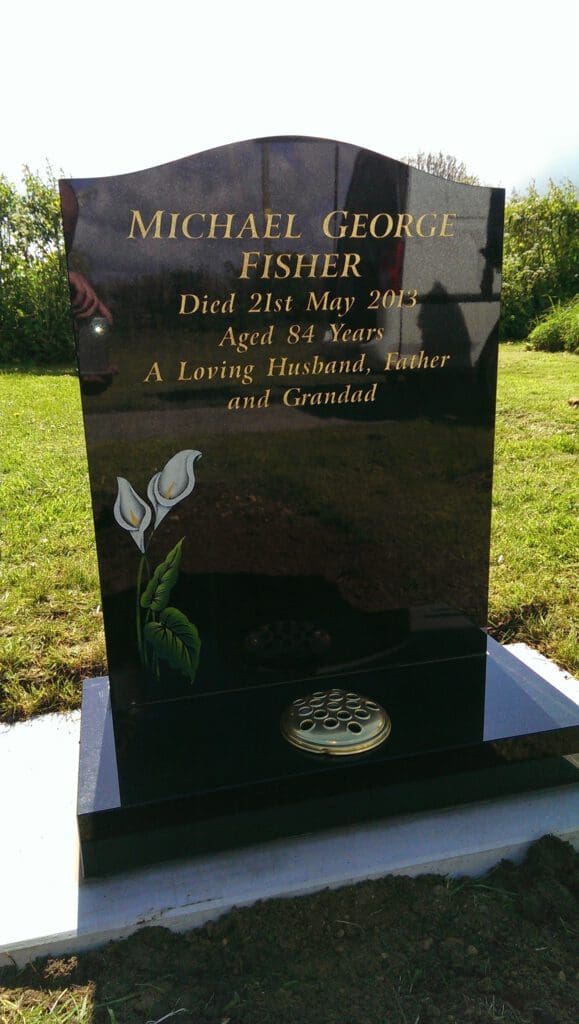 A polished black headstone with gold lettering reads: "Michael George Fisher, Died 21st May 2013, Aged 84 Years, A Loving Husband, Father and Grandad." It also features two white lilies and a brass flower holder at the base.
