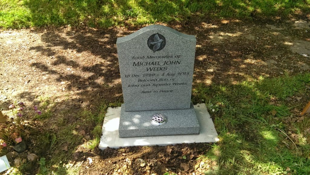 A gravestone for Michael John Weeks, born 16 Dec 1990, died 4 July 2013. It reads "Beloved son of John and Deborah Weeks" and "Rest in Peace." The stone is set on a concrete base surrounded by grass and soil with flowers nearby.
