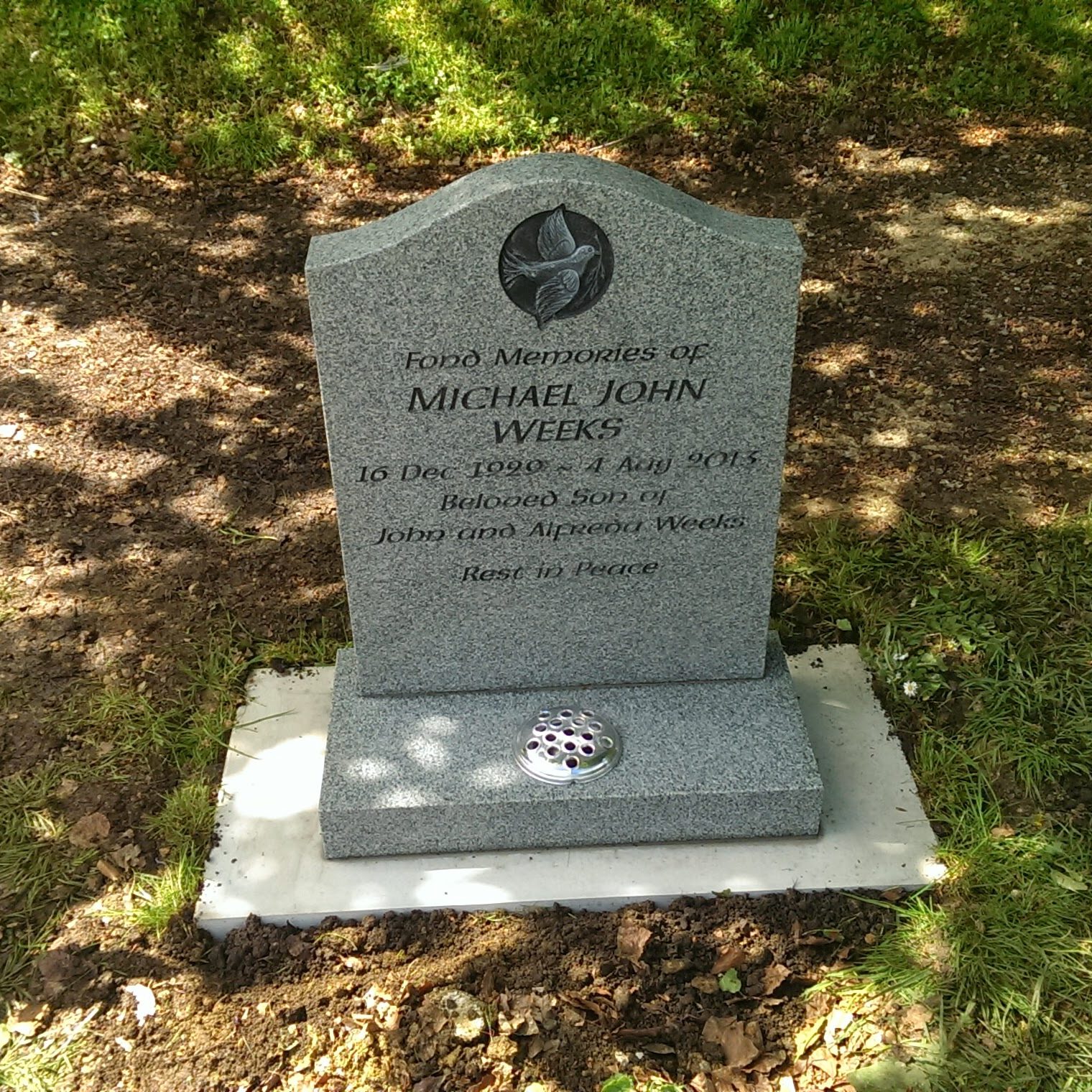 A gravestone for Michael John Weeks, born 16 Dec 1990, died 4 July 2013. It reads "Beloved son of John and Deborah Weeks" and "Rest in Peace." The stone is set on a concrete base surrounded by grass and soil with flowers nearby.