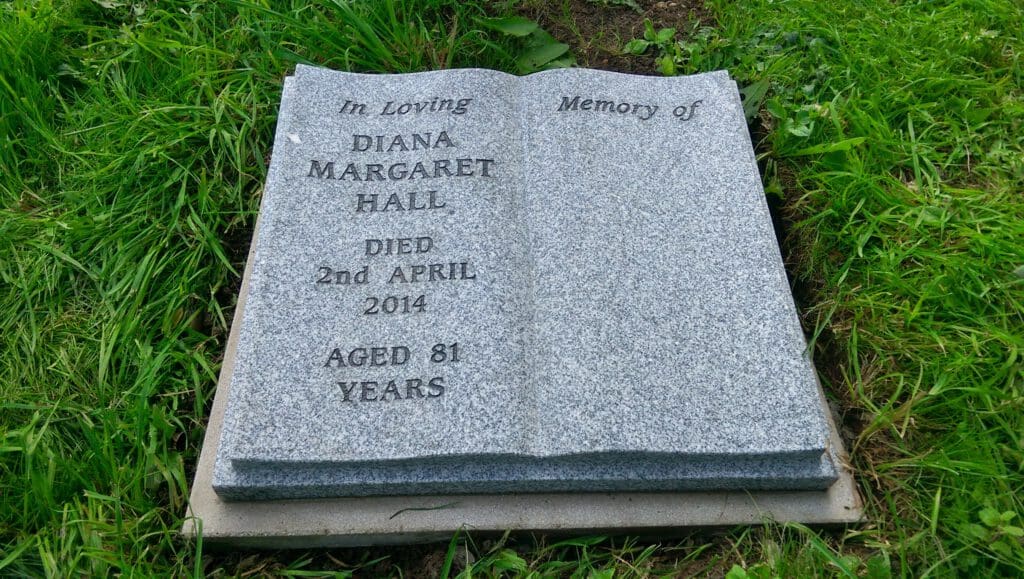 A gray tombstone shaped like an open book lies on green grass. It reads: "In Loving Memory of Diana Margaret Hall. Died 2nd April 2014. Aged 81 Years.