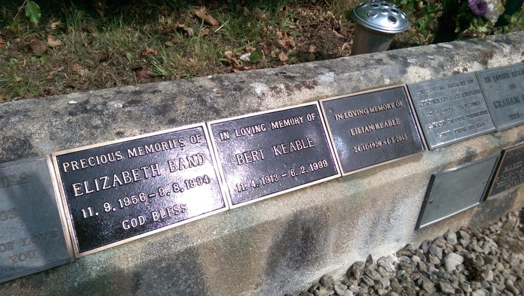 A stone memorial wall with several plaques dedicated to different individuals. Each plaque has engraved names and dates, surrounded by gravel and grass. A silver vase with holes stands in the background.