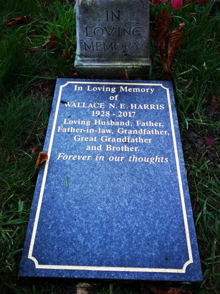 A gravestone with the text: "In Loving Memory of Wallace N.E. Harris 1928-2017. Loving Husband, Father, Father-in-law, Grandfather, Great Grandfather and Brother. Forever in our thoughts." The stone is set on green grass.
