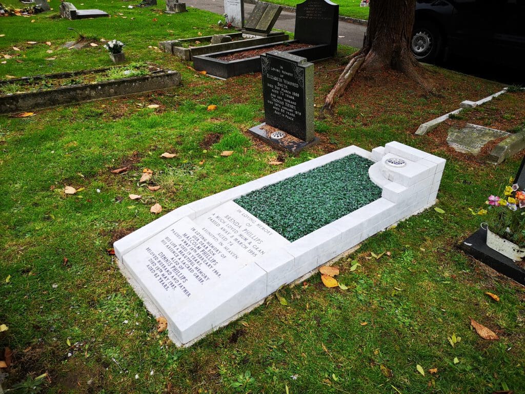 A white marble headstone lies flat on a grassy area, inscribed with names and dates. It has a decorative green stone section and a small raised circular feature at the top. Nearby, an upright black headstone is visible with more inscriptions.