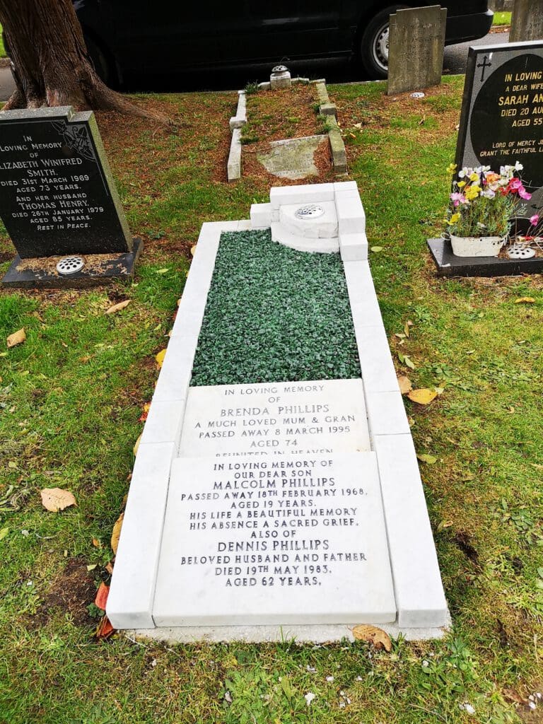 A white marble gravestone with inscriptions, surrounded by greenery. It honors Brenda Phillips, who passed in 1995 at 74, and Malcolm Phillips, who passed in 1968 at 37. Dennis Phillips, who died in 1935 at 62, is also mentioned.