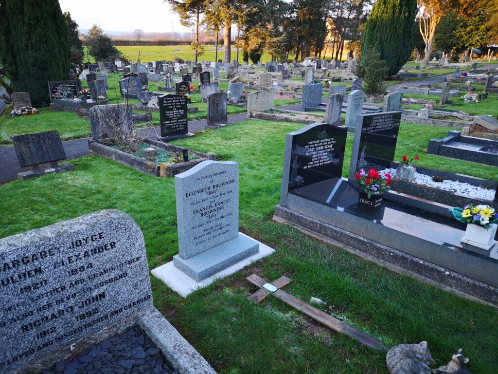 A cemetery with numerous headstones and tombs. Some are engraved with names and dates, while others have flowers or crosses placed nearby. Trees are in the background, and the grass is well-kept.