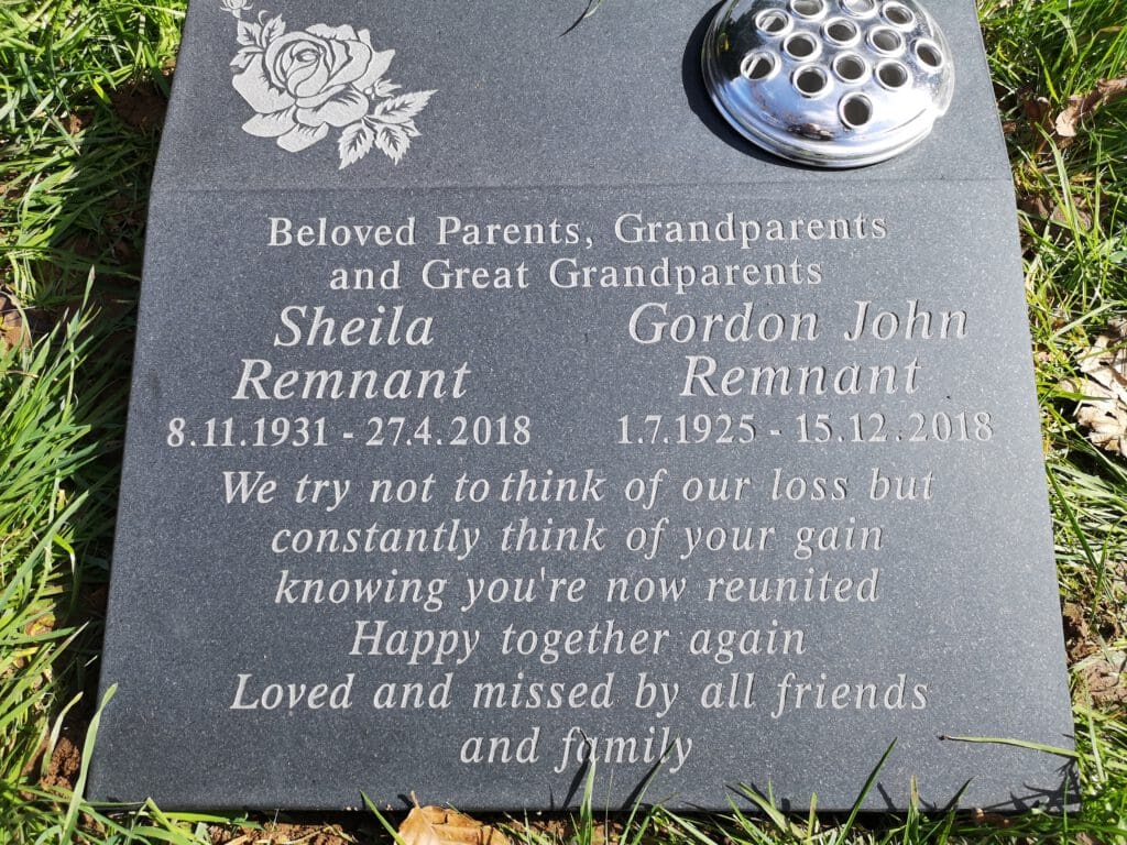A black granite memorial plaque engraved with the names Sheila Remnant and Gordon John Remnant, their birth and death dates, and a sentimental message about loss and reunion. It features a rose design and a metal flower holder.