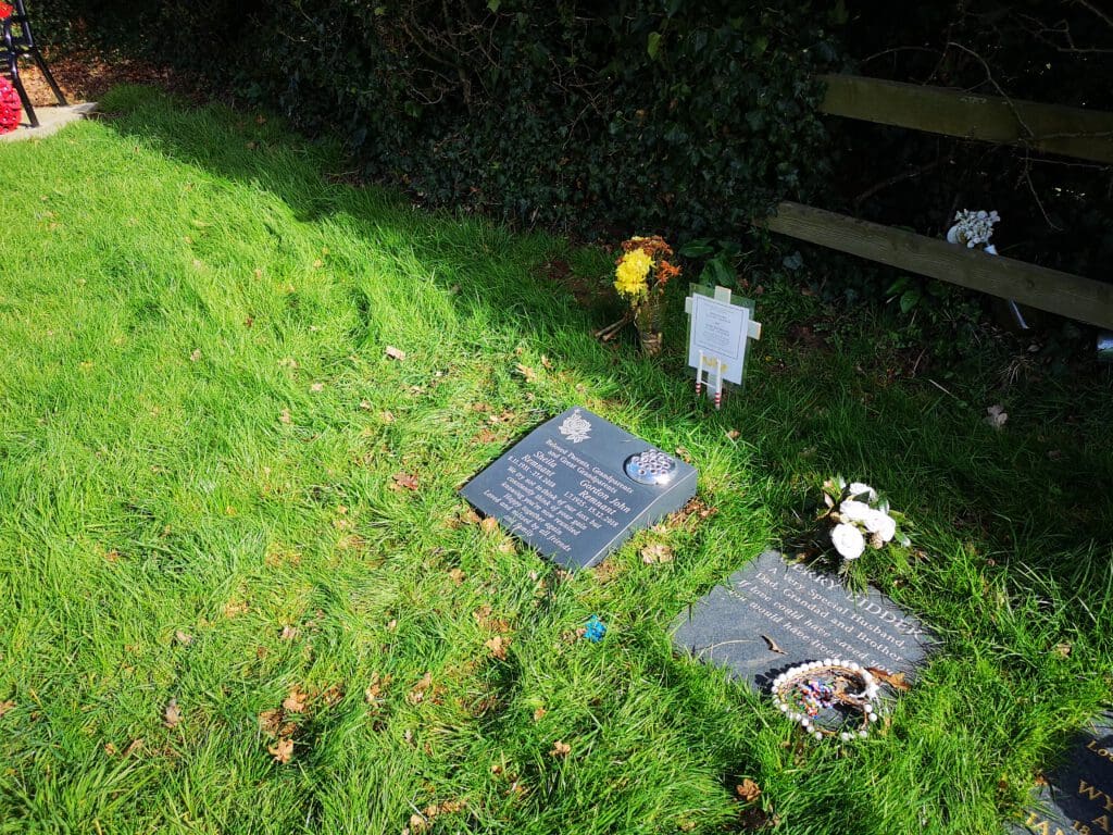 A peaceful cemetery scene shows two graves with plaques embedded in the grass. One plaque is decorated with flowers and small ornamental items, while the other has white flowers and a cross nearby. The ground is covered with vibrant green grass.