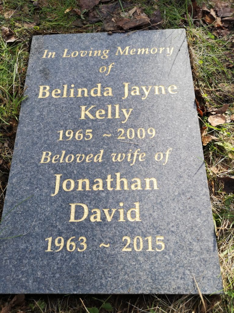 A gravestone on grass reads: "In Loving Memory of Belinda Jayne Kelly, 1965 - 2009, Beloved wife of Jonathan David, 1963 - 2015." Dry leaves are scattered around the stone.