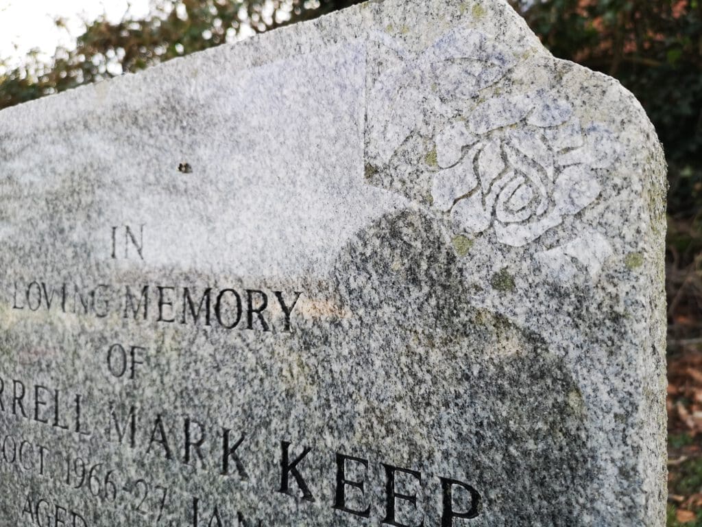A close-up of a weathered, gray tombstone with engraved text partially visible, reading "In Loving Memory." The stone's corner features a carved rose design. The background shows blurred greenery.