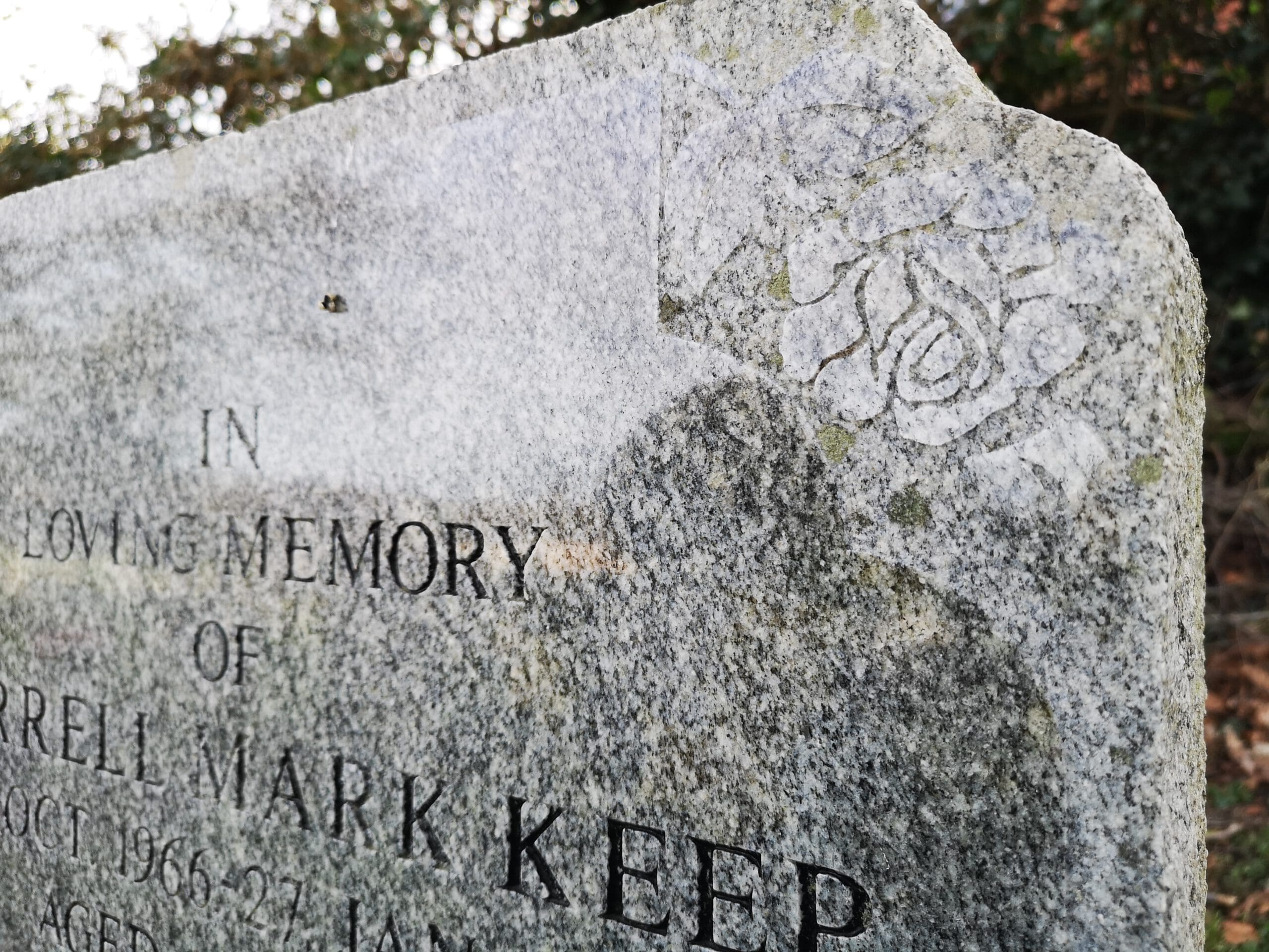 A close-up of a weathered, gray tombstone with engraved text partially visible, reading "In Loving Memory." The stone's corner features a carved rose design. The background shows blurred greenery.