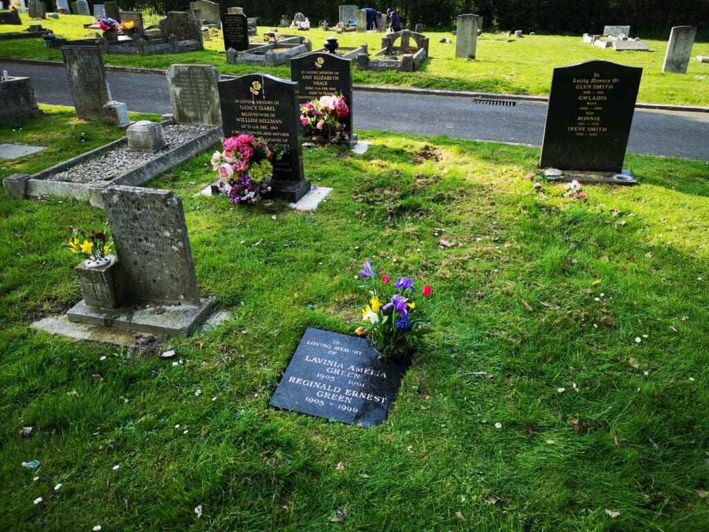 A cemetery scene showing several gravestones with colorful flower arrangements. The graves are set on a green lawn. One stone in the foreground is horizontal, while others in the background are upright. A paved path runs behind the graves.
