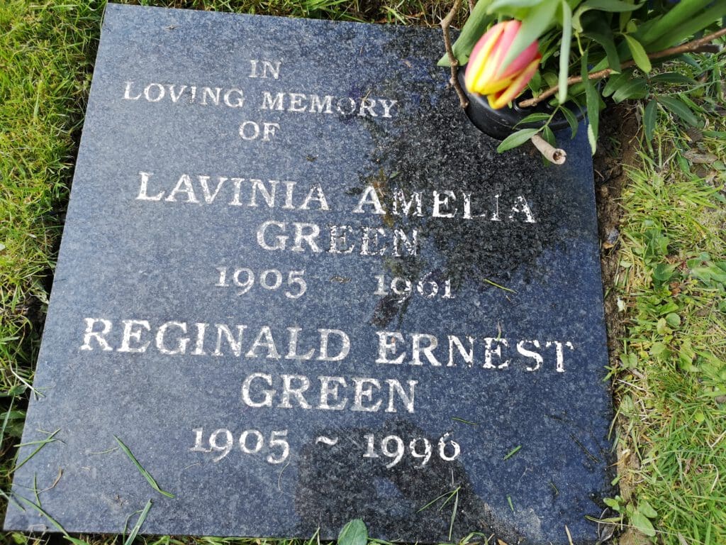 A black gravestone reads: "In Loving Memory of Lavinia Amelia Green 1905-1991, Reginald Ernest Green 1905-1996." A tulip is placed on the top right corner, surrounded by grass.