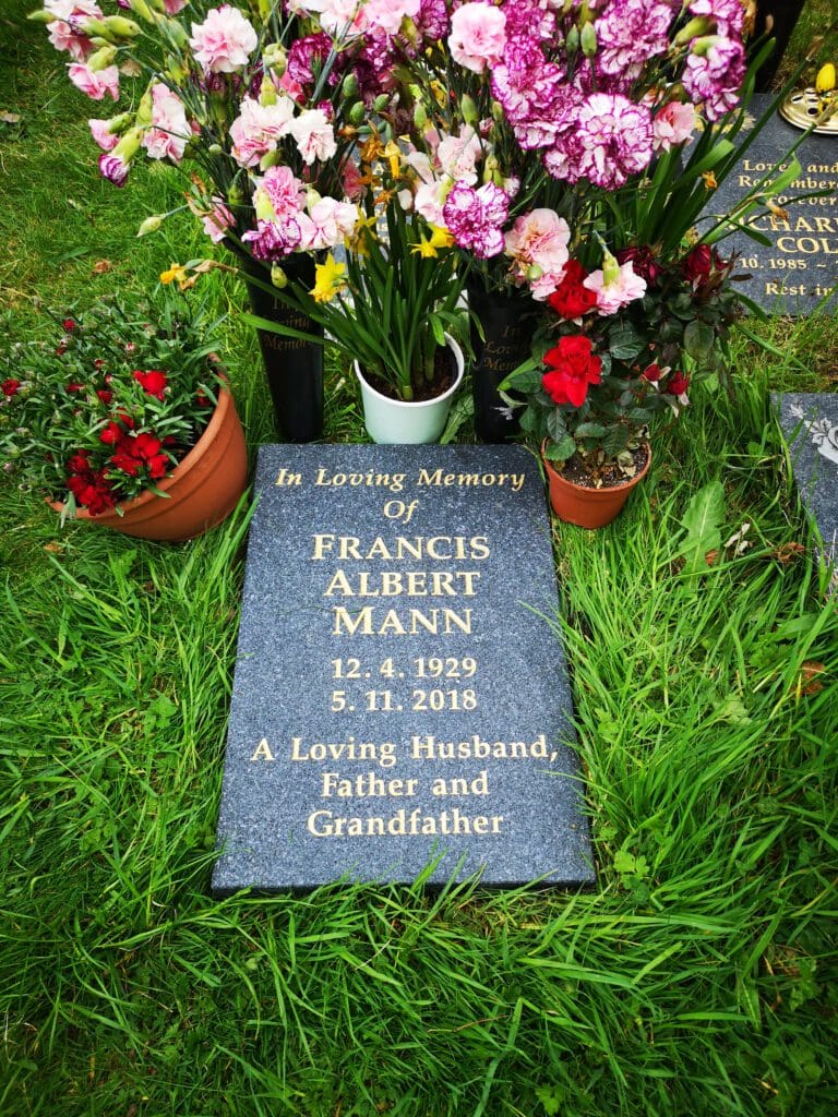 A tombstone on grass reads "In Loving Memory of Francis Albert Mann, 12.4.1929 - 5.11.2018, A Loving Husband, Father and Grandfather." It's surrounded by colorful flowers in vases and pots.