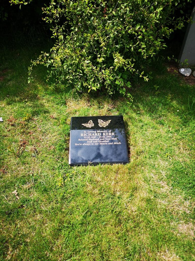 A black grave marker on lush green grass, partially shaded by a small bush. The marker displays two butterfly designs above the engraved text. The surrounding area is serene, highlighting the grave's peaceful setting.