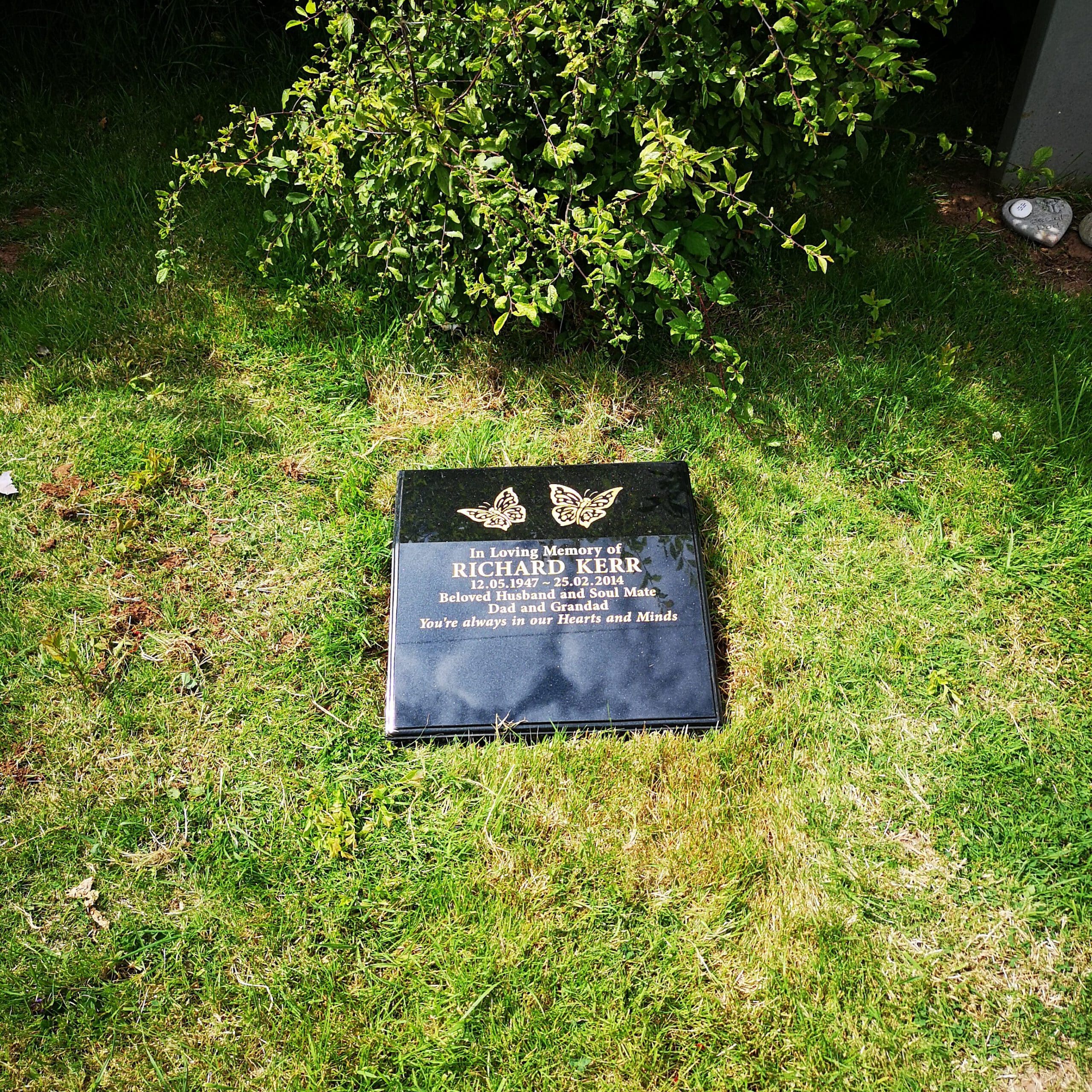 A black grave marker on lush green grass, partially shaded by a small bush. The marker displays two butterfly designs above the engraved text. The surrounding area is serene, highlighting the grave's peaceful setting.