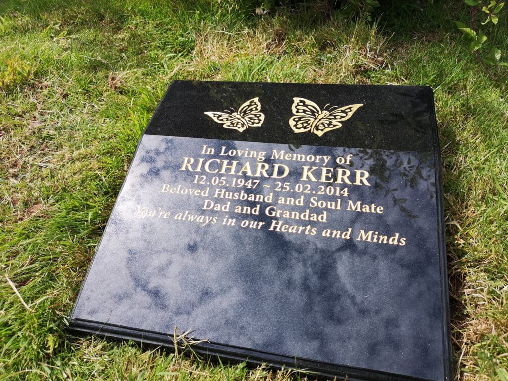A black memorial plaque set in grass, inscribed with "In Loving Memory of Richard Kerr," and dates of birth and death. It features two butterfly motifs and the message, "You're always in our Hearts and Minds.