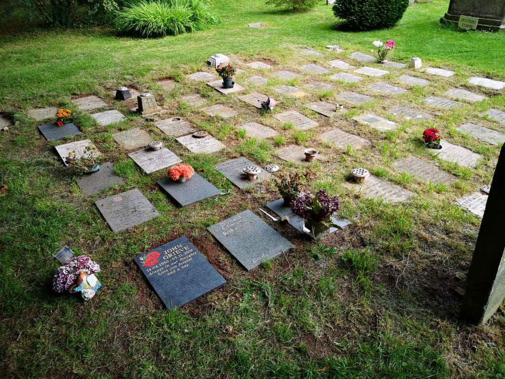 A grassy cemetery area with rows of flat headstones. Some headstones have flowers in pots or small arrangements placed on top. The grass and nearby bushes are lush and green, indicating a well-maintained area.