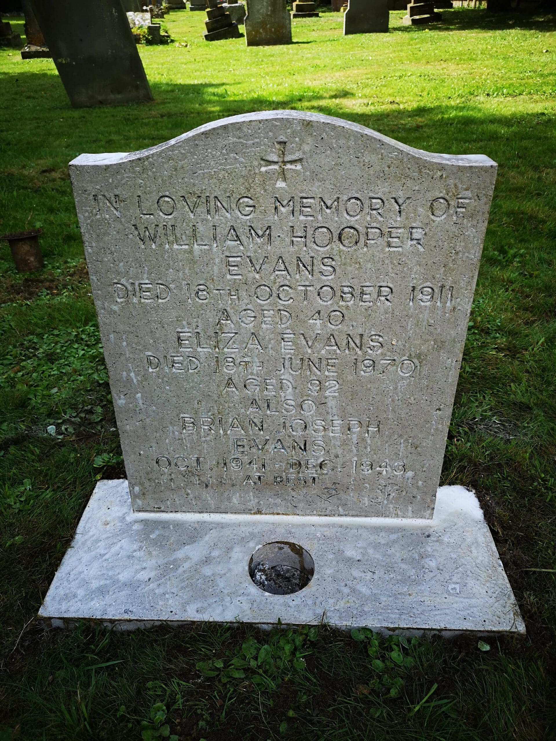 A gravestone in a cemetery with text commemorating William Hooper Evans, Eliza Evans, and Brian Joseph Evans, listing their dates of death and ages. The gravestone is surrounded by grass, with other gravestones in the background.