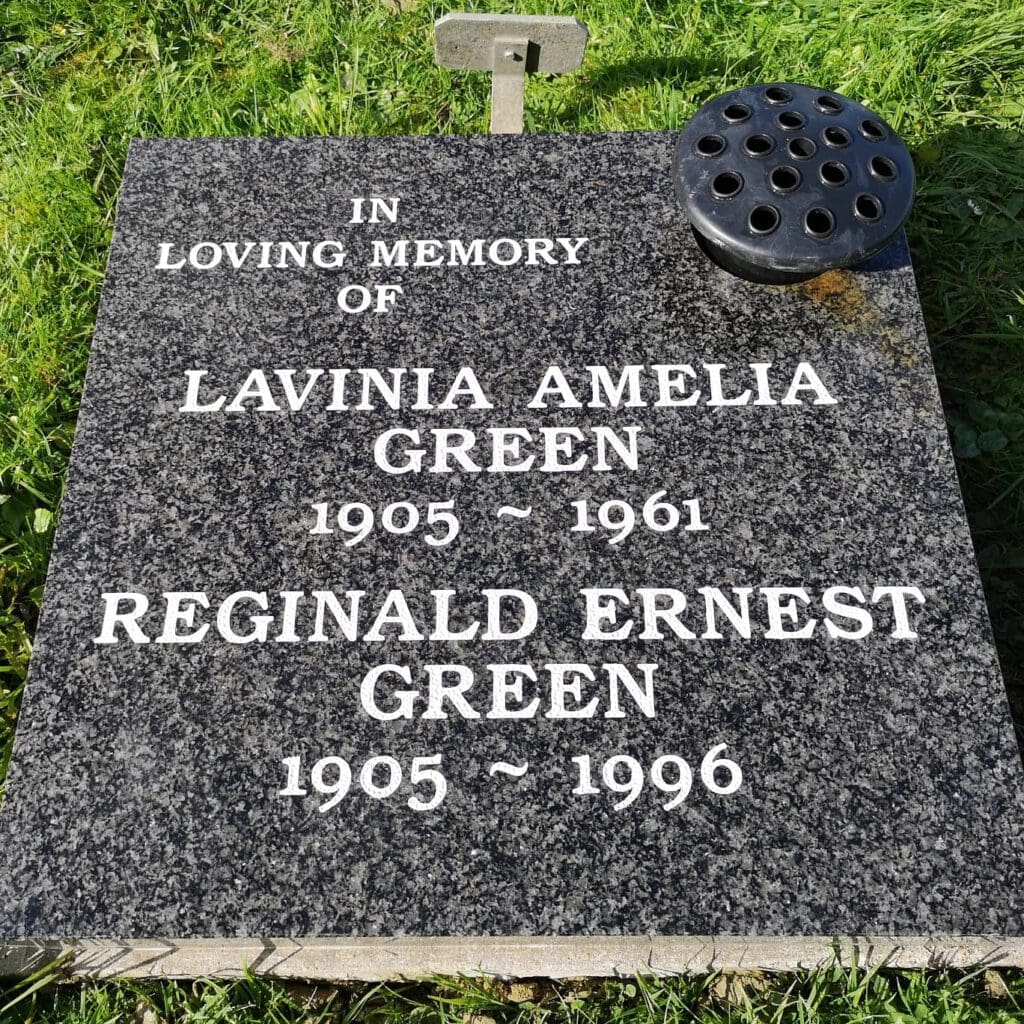 A polished black gravestone on grass reads: "In loving memory of Lavinia Amelia Green 1905 ~ 1961, Reginald Ernest Green 1905 ~ 1996." A circular flower holder is positioned at the top.