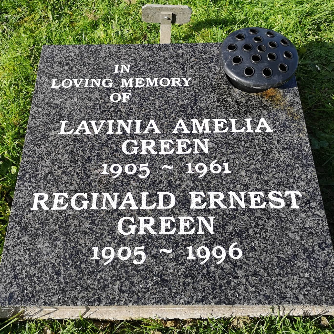 A polished black gravestone on grass reads: "In loving memory of Lavinia Amelia Green 1905 ~ 1961, Reginald Ernest Green 1905 ~ 1996." A circular flower holder is positioned at the top.