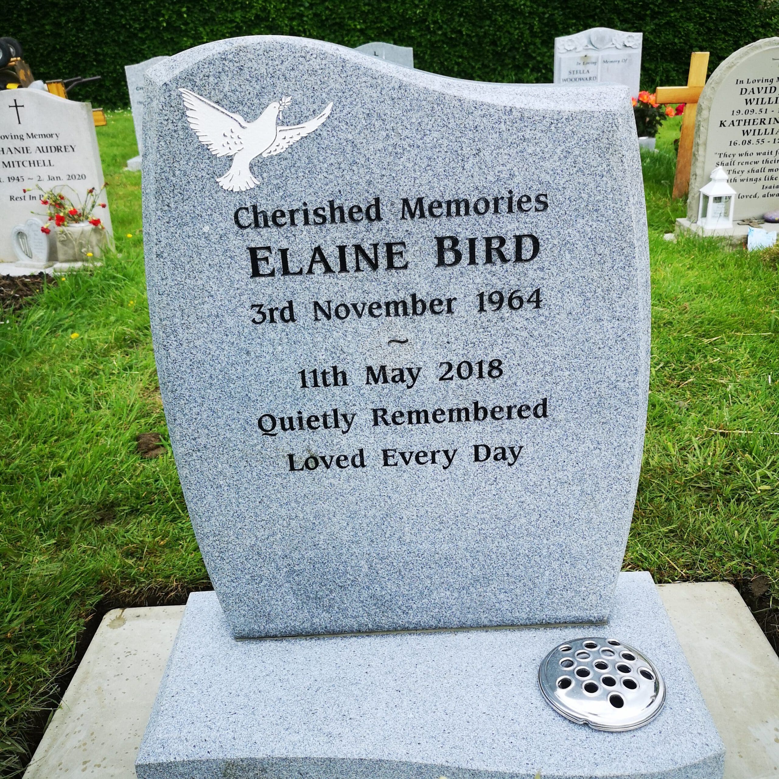 A gray gravestone in a cemetery is inscribed with "Cherished Memories Elaine Bird, 3rd November 1964 - 11th May 2018. Quietly Remembered, Loved Every Day." A white dove symbol is engraved at the top. Flowers and other graves are in the background.