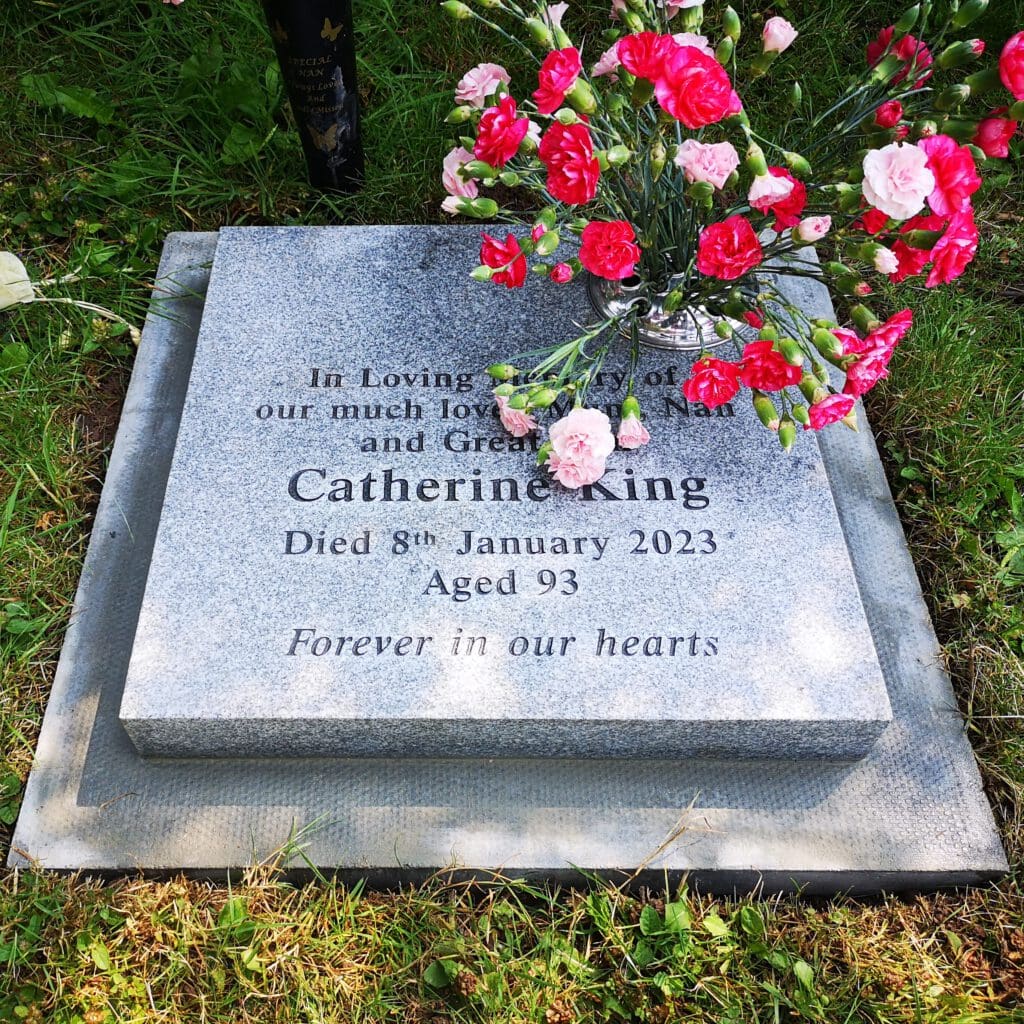 A gravestone in a grassy area with flowers on top. The inscription reads: "In Loving Memory of our much loved Mum, Nan, and Great Nan, Catherine King. Died 8th January 2023 Aged 93. Forever in our hearts." Colorful flowers are laid around it.