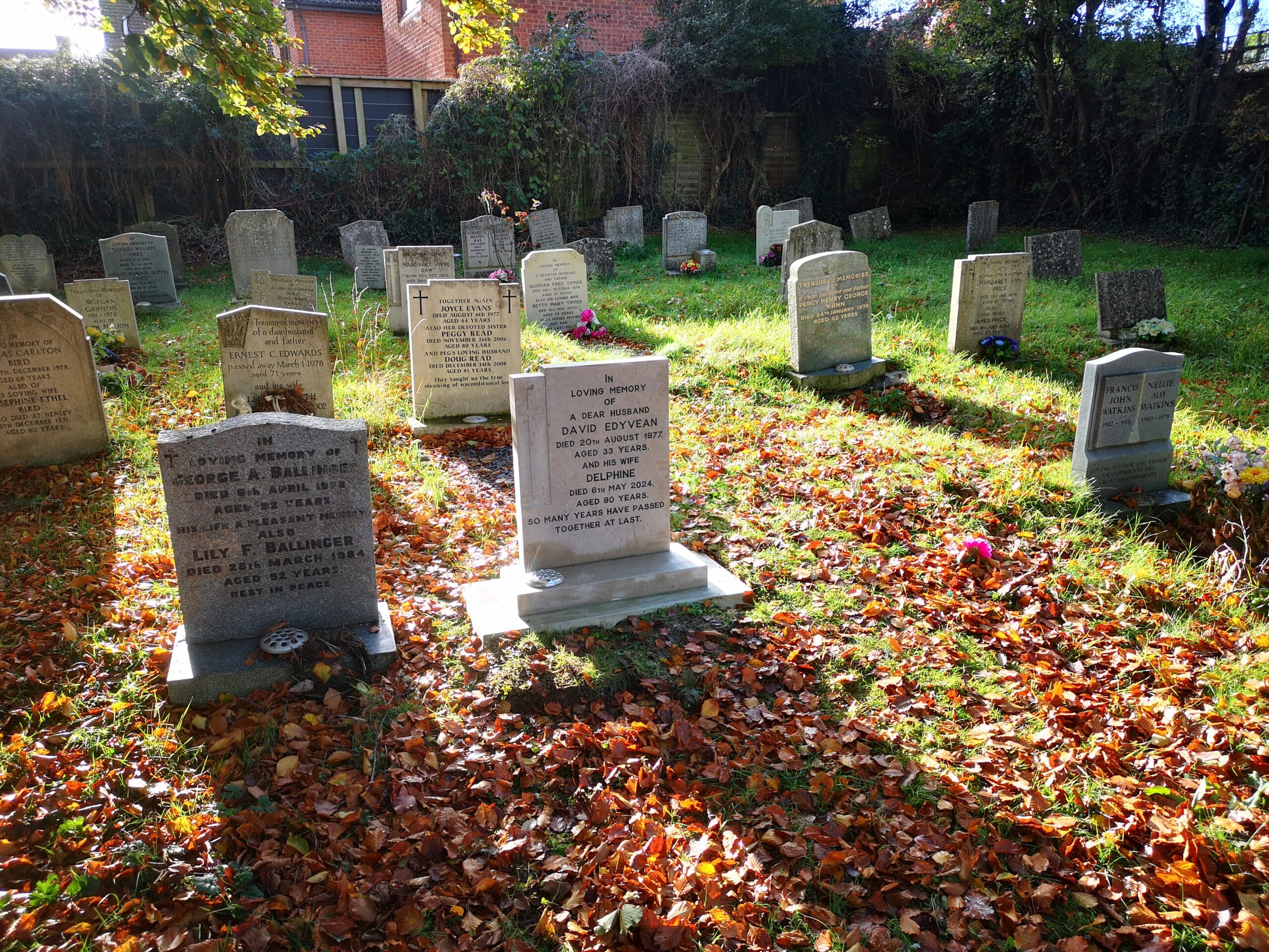 A small cemetery with several headstones surrounded by fallen autumn leaves. The sunlight creates patches of light on the ground, highlighting the stones and a few flower arrangements placed in remembrance. Trees and a building are in the background.