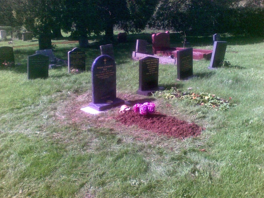 A cemetery with several headstones, including a newly filled grave with dark soil and a small bouquet of bright pink flowers placed on top. The area is surrounded by green grass and trees in the background.