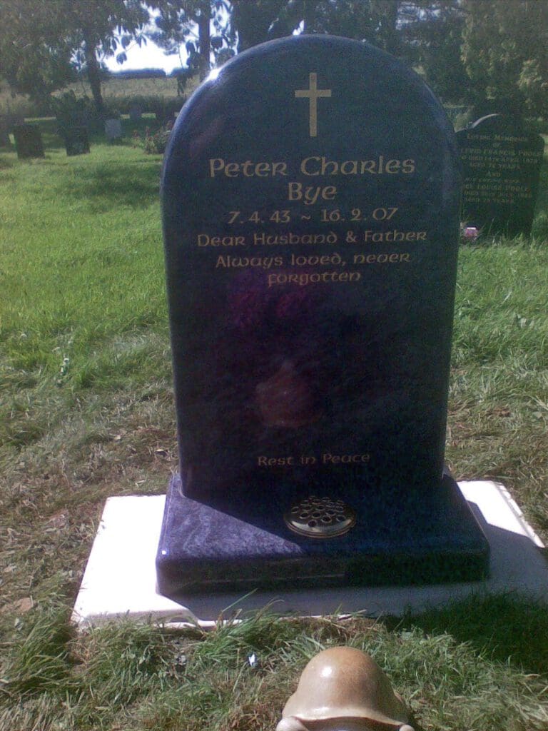 A gravestone in a grassy cemetery. It reads, "Peter Charles Bye, 7.4.43 - 16.2.07. Dear Husband & Father, Always loved, never forgotten. Rest in Peace." A cross is engraved at the top.