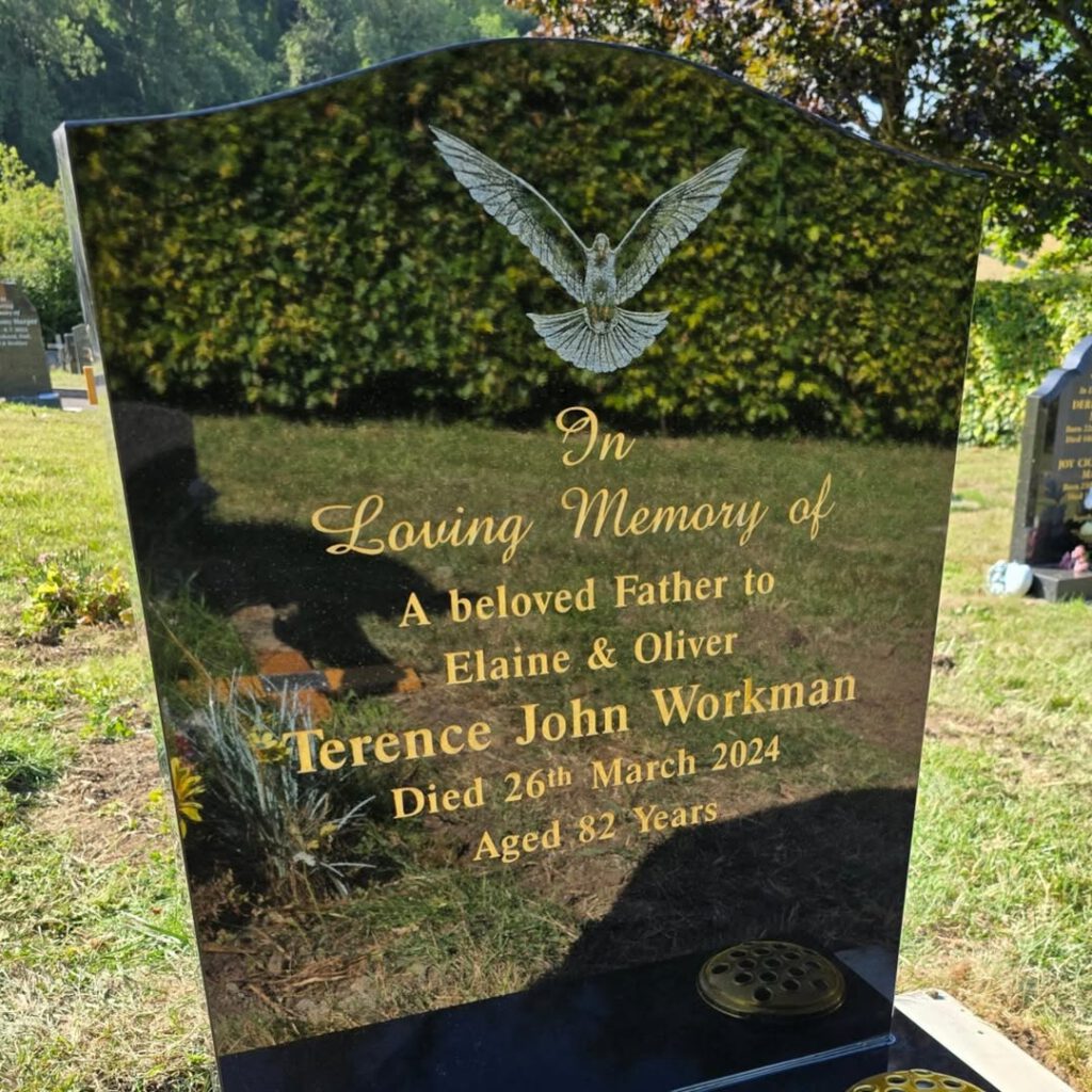 A polished black headstone with a carved dove and gold inscription reads: "In Loving Memory of Terence John Workman, beloved father to Elaine & Oliver, died 26th March 2024, aged 82 years.