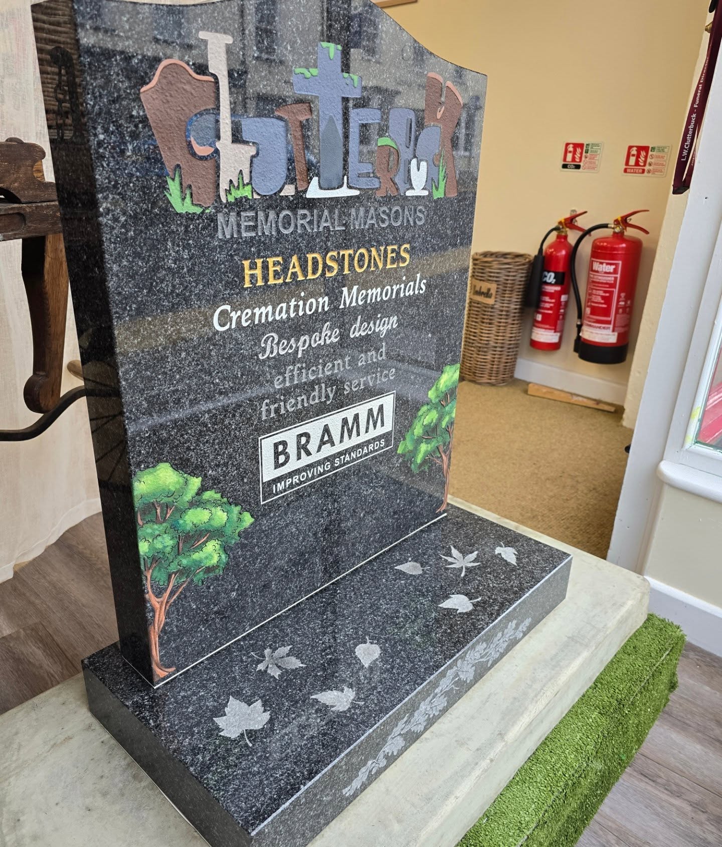 A polished black headstone with colorful lettering advertises memorial masonry services, including headstones and cremation memorials. The base features engraved leaves, and fire extinguishers are seen in the background.