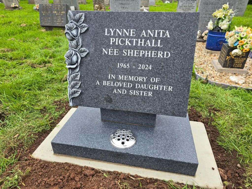 A gravestone with rose carvings reads: "Lynne Anita Pickthall, née Shepherd, 1965-2024. In memory of a beloved daughter and sister." It stands on grass among other graves and a pot of flowers.