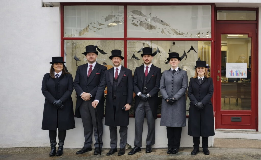 Six people dressed in formal funeral attire, including black coats, gloves, and top hats, stand in a row outside a building with a red-framed window and door. The atmosphere is professional and solemn.