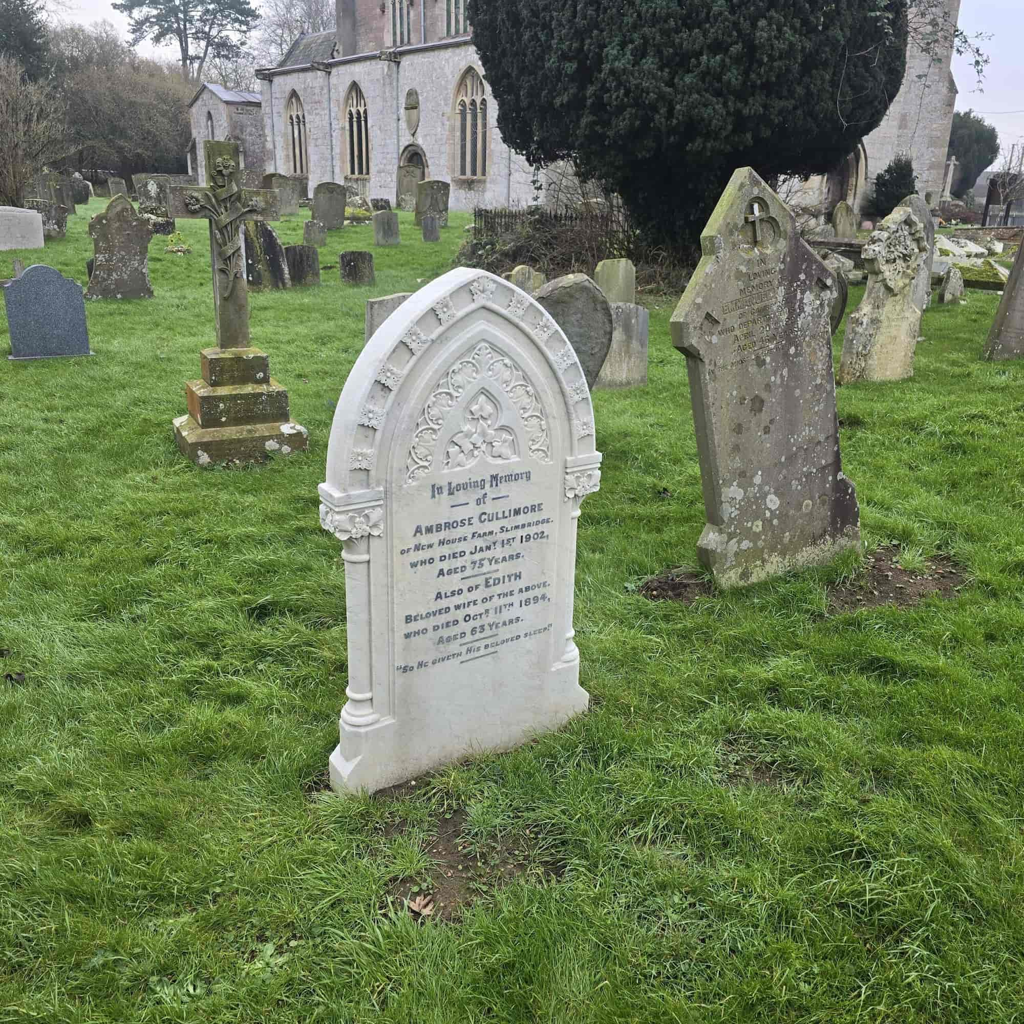 A white gravestone with engraved text stands in a grassy cemetery, surrounded by older, weathered headstones. A stone church with arched windows is visible in the background amid several trees.