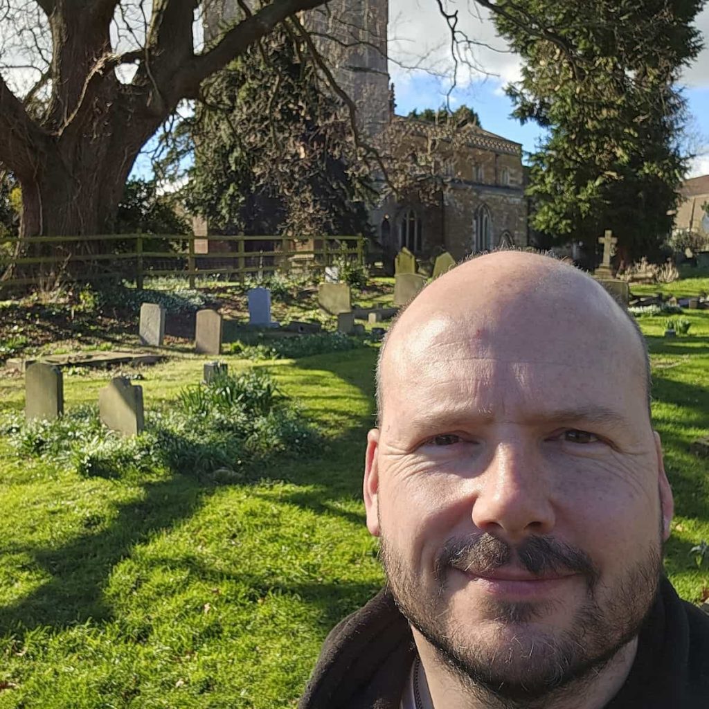 A man with a bald head and beard smiles in the foreground of a sunny graveyard, with gravestones, grass, a large tree, and an old church building visible behind him.