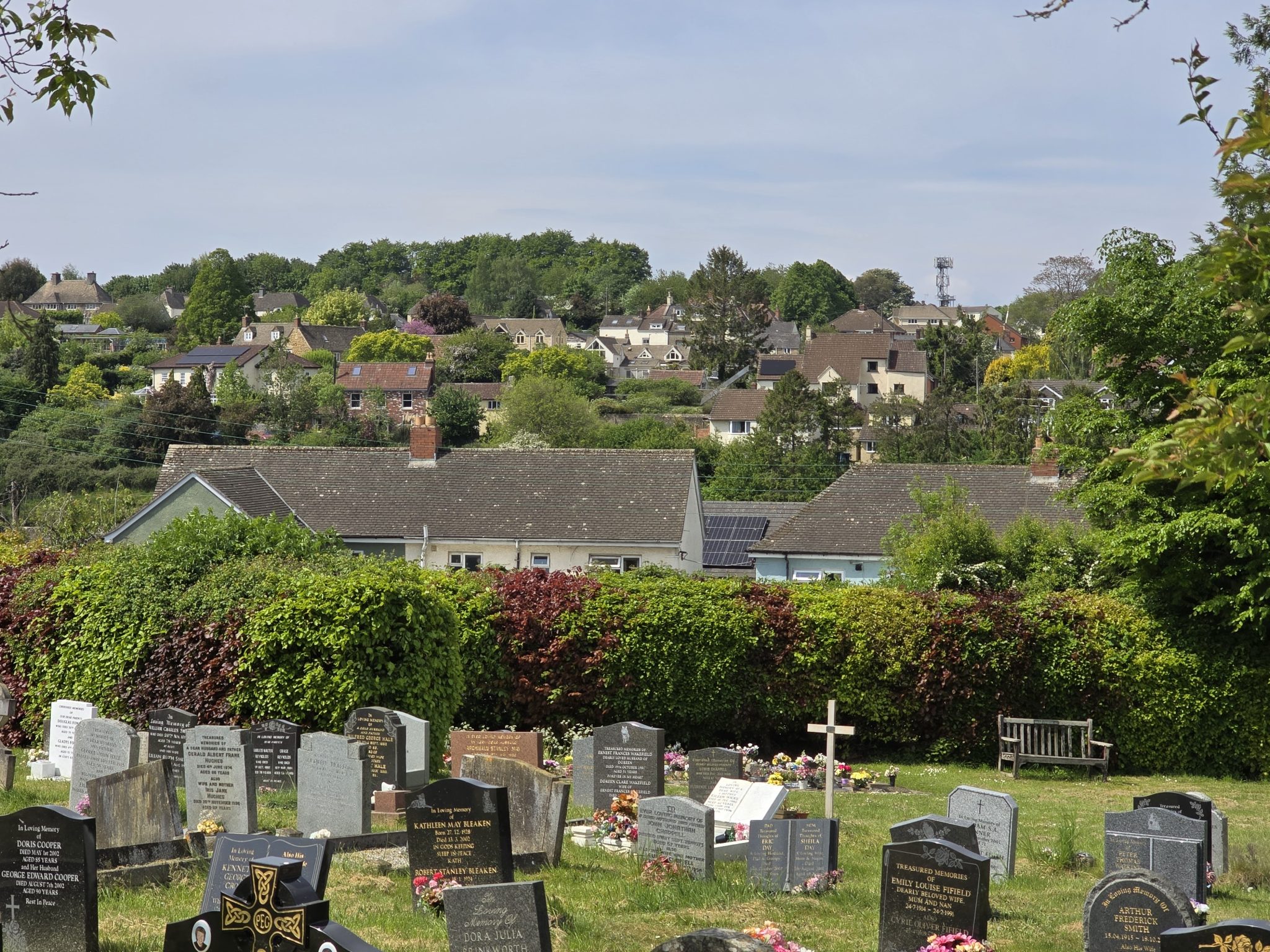 A cemetery with headstones and flowers in the foreground, a bench nearby, and a background of houses, trees, and a hilly landscape under a partly cloudy sky.