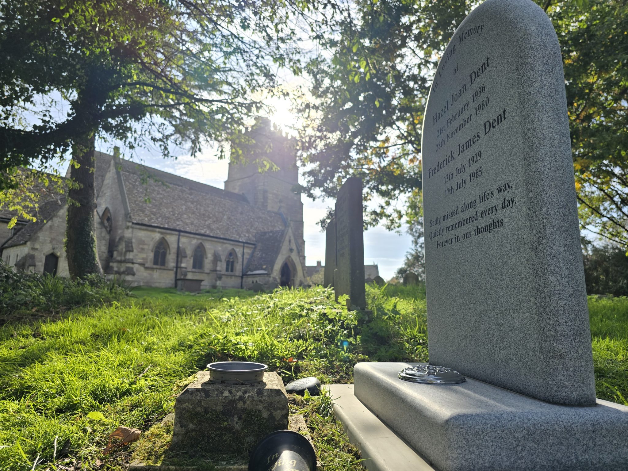 A gravestone stands in a sunlit churchyard with an old stone church in the background, surrounded by trees and grass.