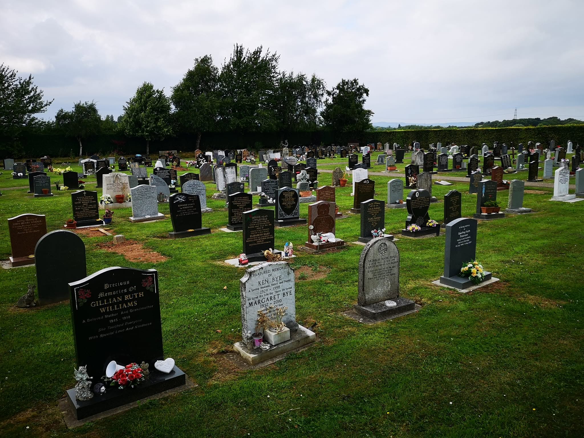 A large cemetery with rows of gravestones on green grass, some decorated with flowers and wreaths, under a cloudy sky. Trees line the background.