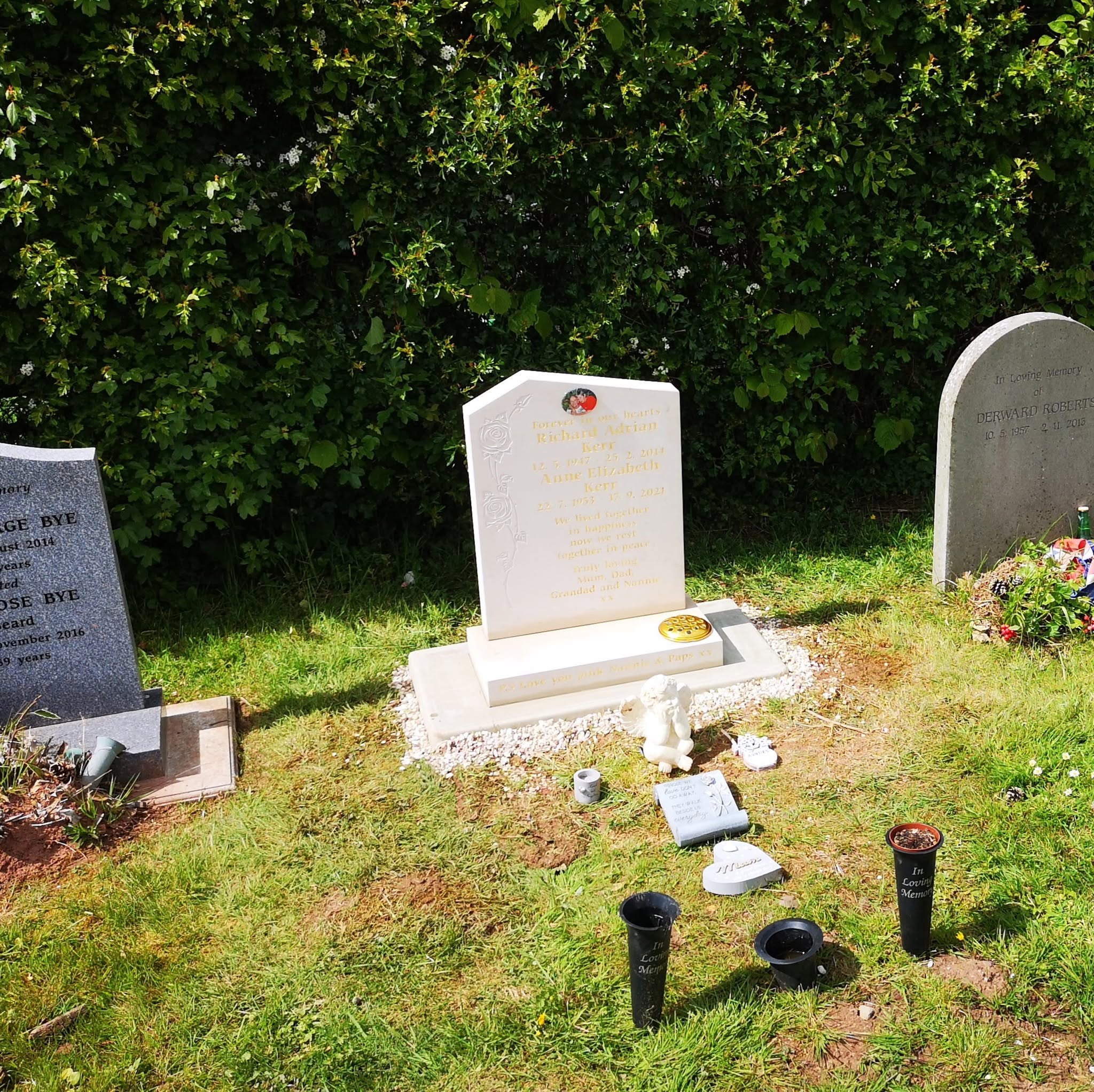 A white gravestone with tribute items in front stands on grass, flanked by two other gravestones. The background is dense green foliage, and sunlight casts shadows on the scene.