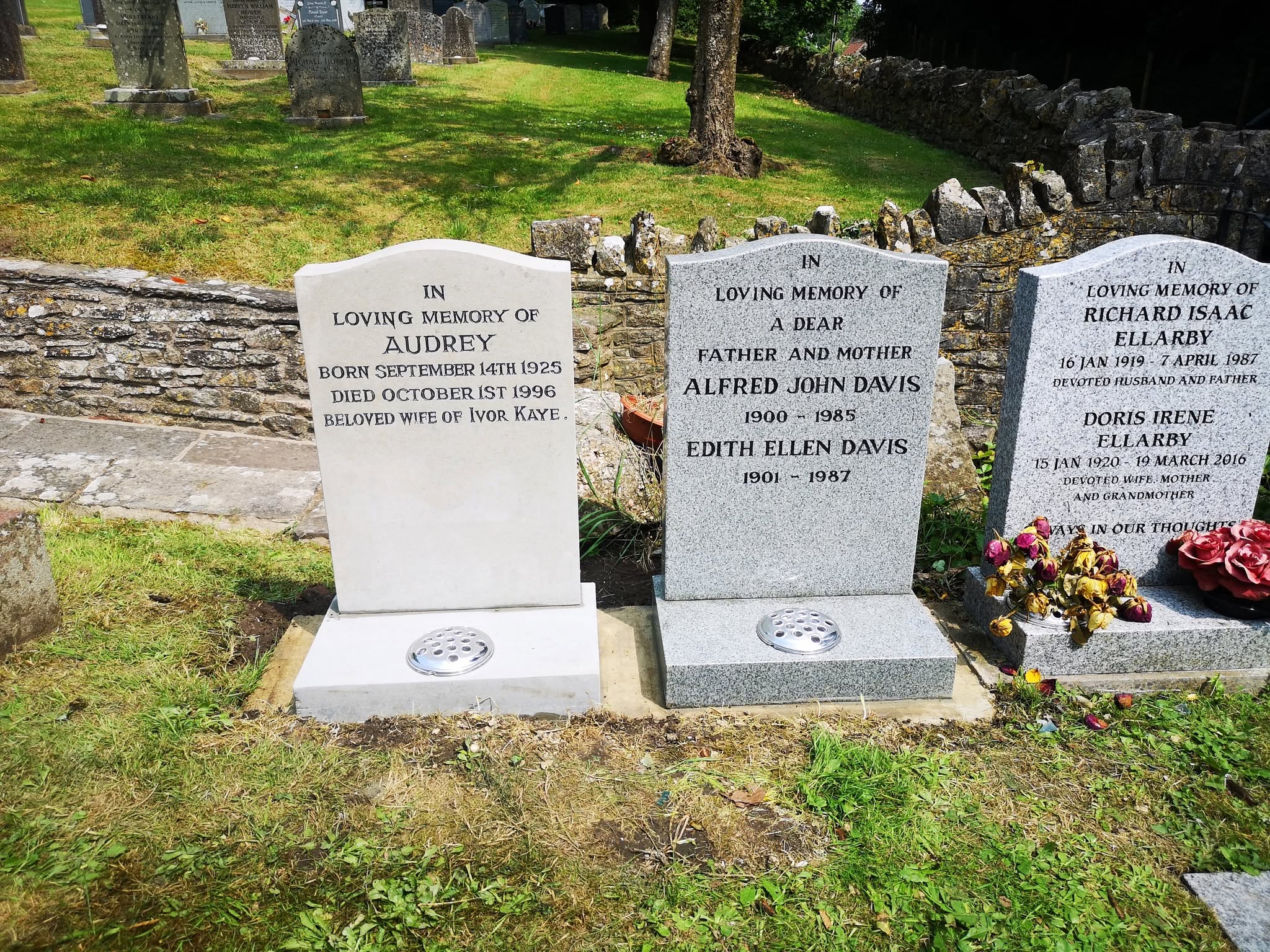 Three gravestones in a cemetery, each dedicated to different individuals. The middle grave has fresh flowers. The stones are set on green grass, with a stone wall and more gravestones visible in the background.