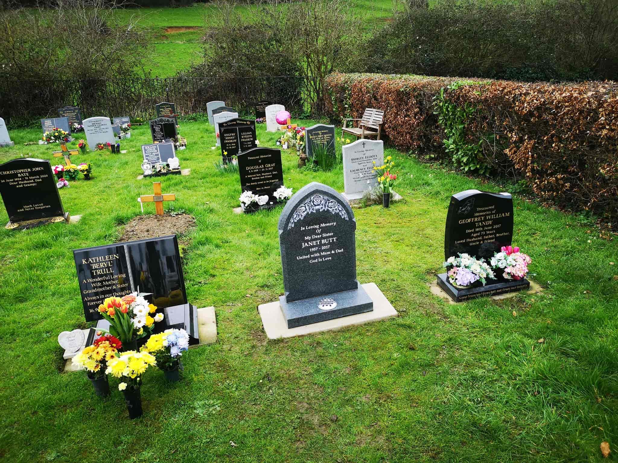 A cemetery with several gravestones and flower arrangements on green grass, surrounded by bushes and trees in the background.