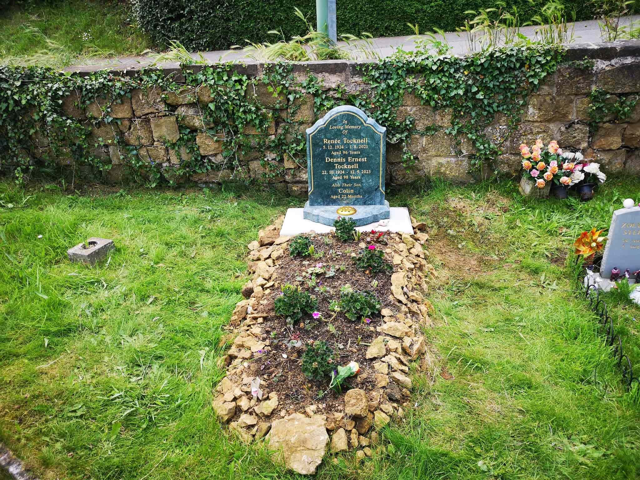 A gravestone with inscriptions stands at the head of a grave outlined with stones and small plants, set in a grassy area near a stone wall and a bouquet of artificial flowers.