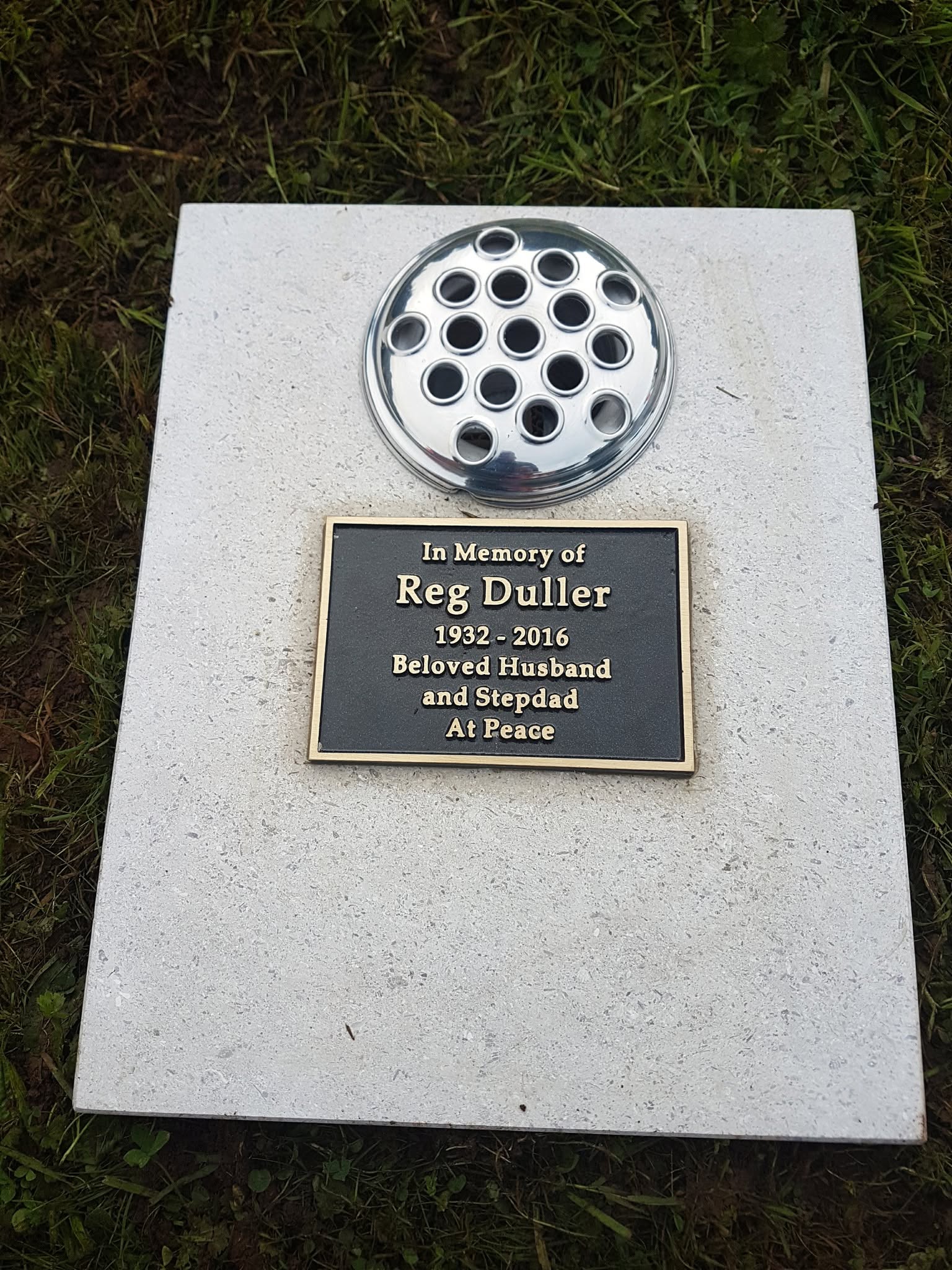 A gravestone plaque reads: "In Memory of Reg Duller 1932-2016 Beloved Husband and Stepdad At Peace." A circular flower holder is above the plaque, and the stone rests on grass.