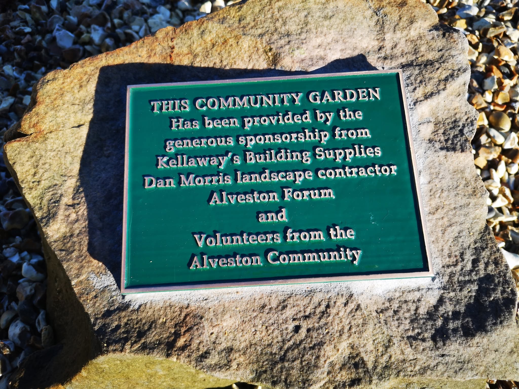 A green plaque on a stone reads: "This community garden has been provided by sponsorship from Kellaway's Building Supplies, Dan Morris, Alveston Forum, and volunteers from the Alveston community.