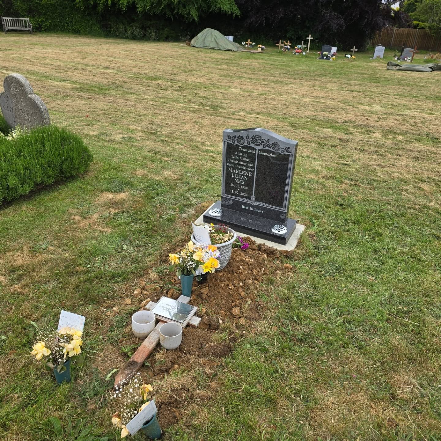 A grave with a black headstone, fresh soil, flowers, small pots, and a spade in a grassy cemetery with other graves and a green tent visible in the background.