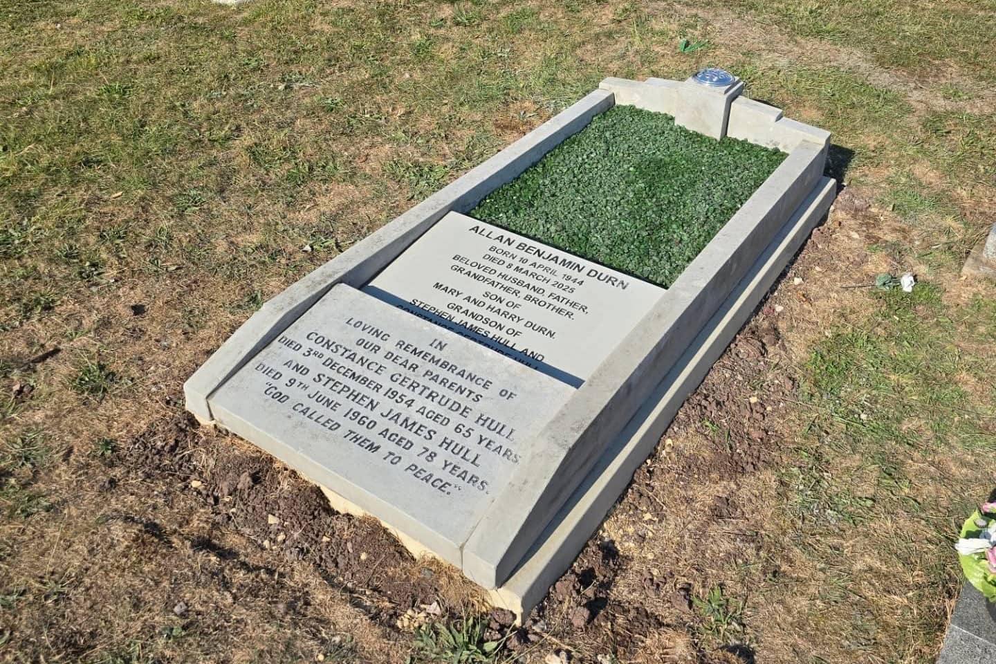 A gravestone in a grassy cemetery with inscriptions for Allan Benjamin Dunn and family members, set in a concrete border, with artificial grass covering the grave plot.