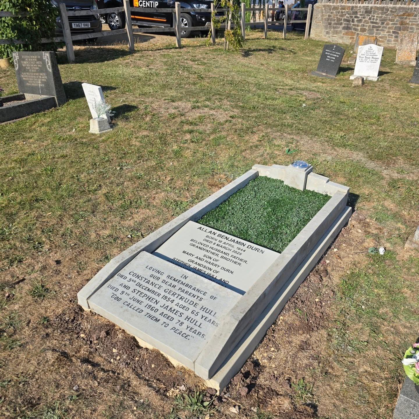 A grave with inscriptions in English, surrounded by grass. The main headstone reads “Allan Benjamin Durn, Loving Dad, Grandad” and includes dates and a tribute. Several other gravestones are visible in the background.