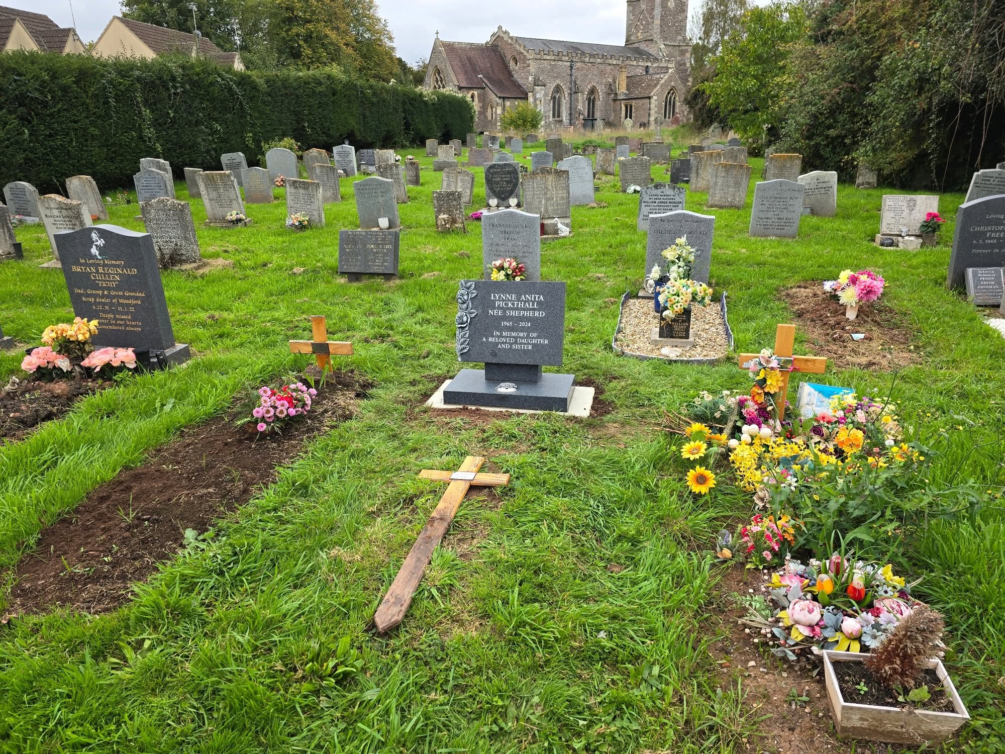 A cemetery with headstones, crosses, and colorful flower arrangements on several graves. A church building is visible in the background, surrounded by trees and greenery.
